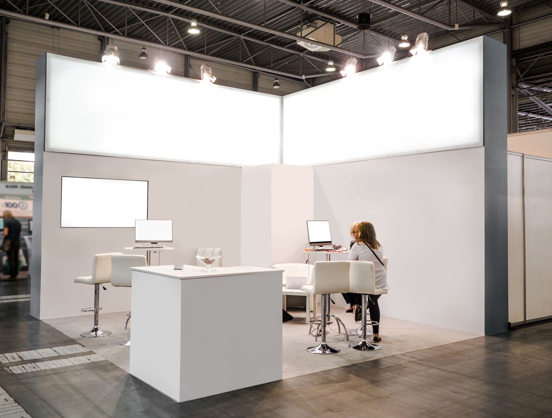 Minimalist white exhibition booth with tables, chairs, and two people seated inside at a trade show.