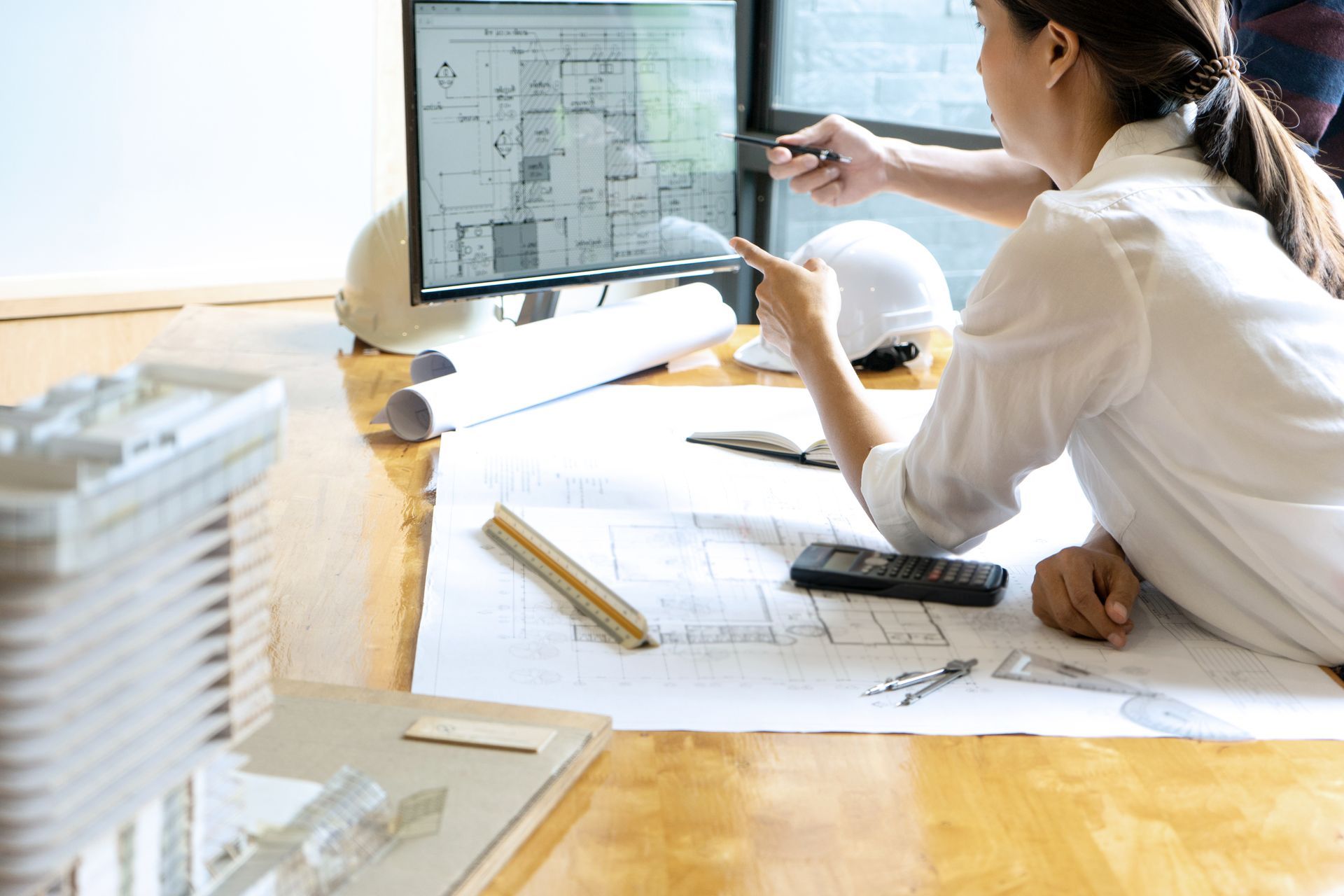 Person reviewing architectural plans at a desk with a monitor displaying floor plans