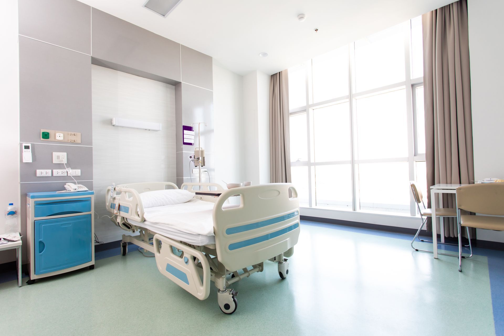 Empty hospital room with a bed, medical equipment, and large windows with curtains Empty hospital room with a bed, medical equipment, and large windows with curtains