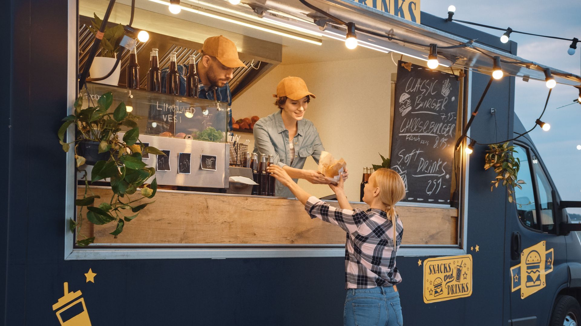 Food truck window with a vendor serving a customer in a denim jacket and yellow cap at dusk