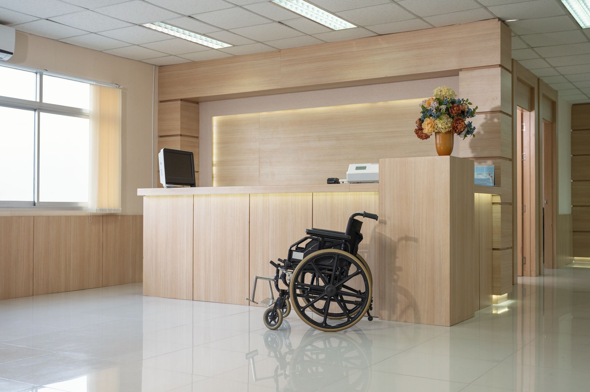 Wheelchair at a modern reception desk in a bright lobby with flowers and a window.