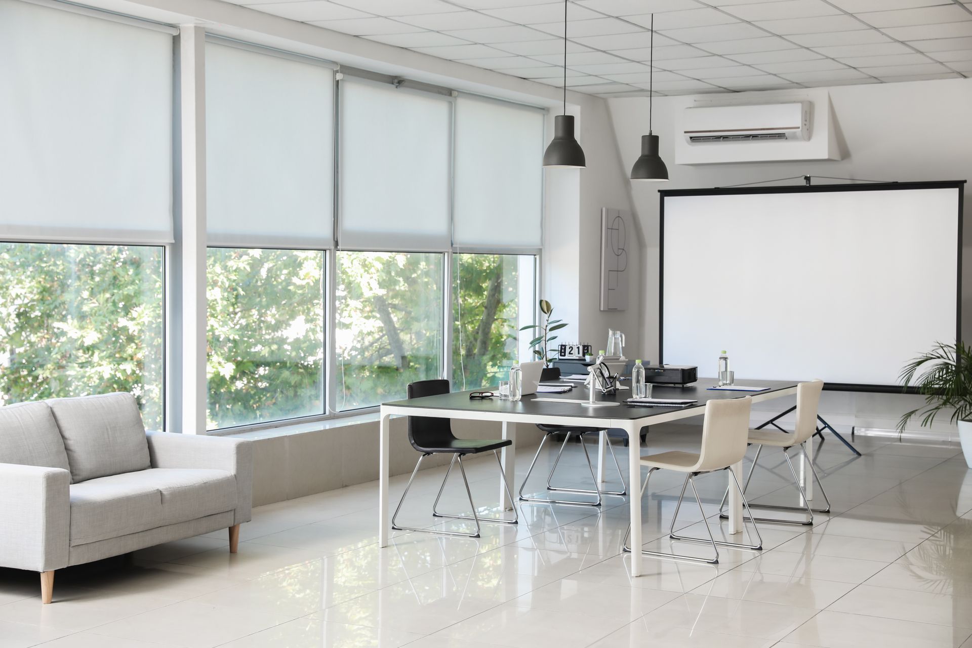 Bright modern meeting room with large windows, white tables, chairs, projector screen, and hanging lights