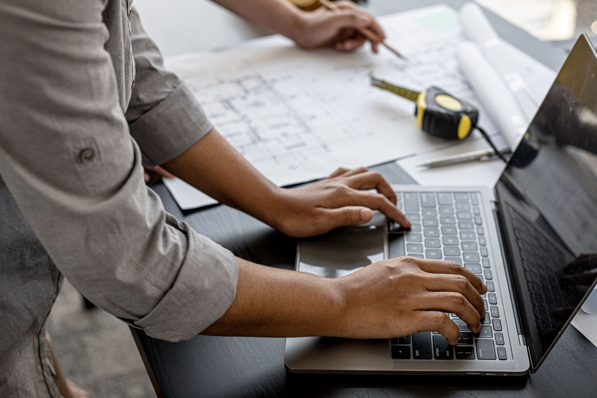 Hands typing on a laptop at a desk with papers and a measuring tape nearby. Hands typing on a laptop at a desk with papers and a measuring tape nearby.