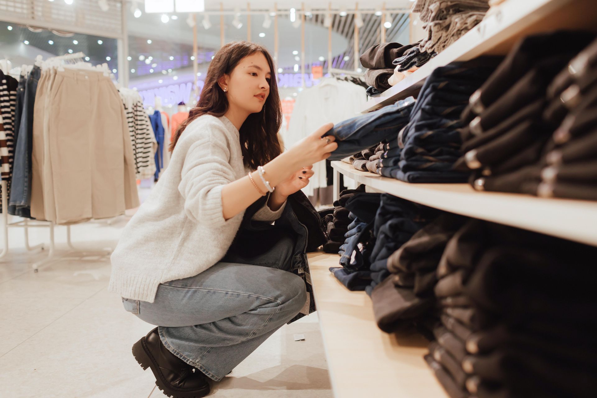 Woman crouches in a clothing store, examining folded black sweaters on a shelf.
