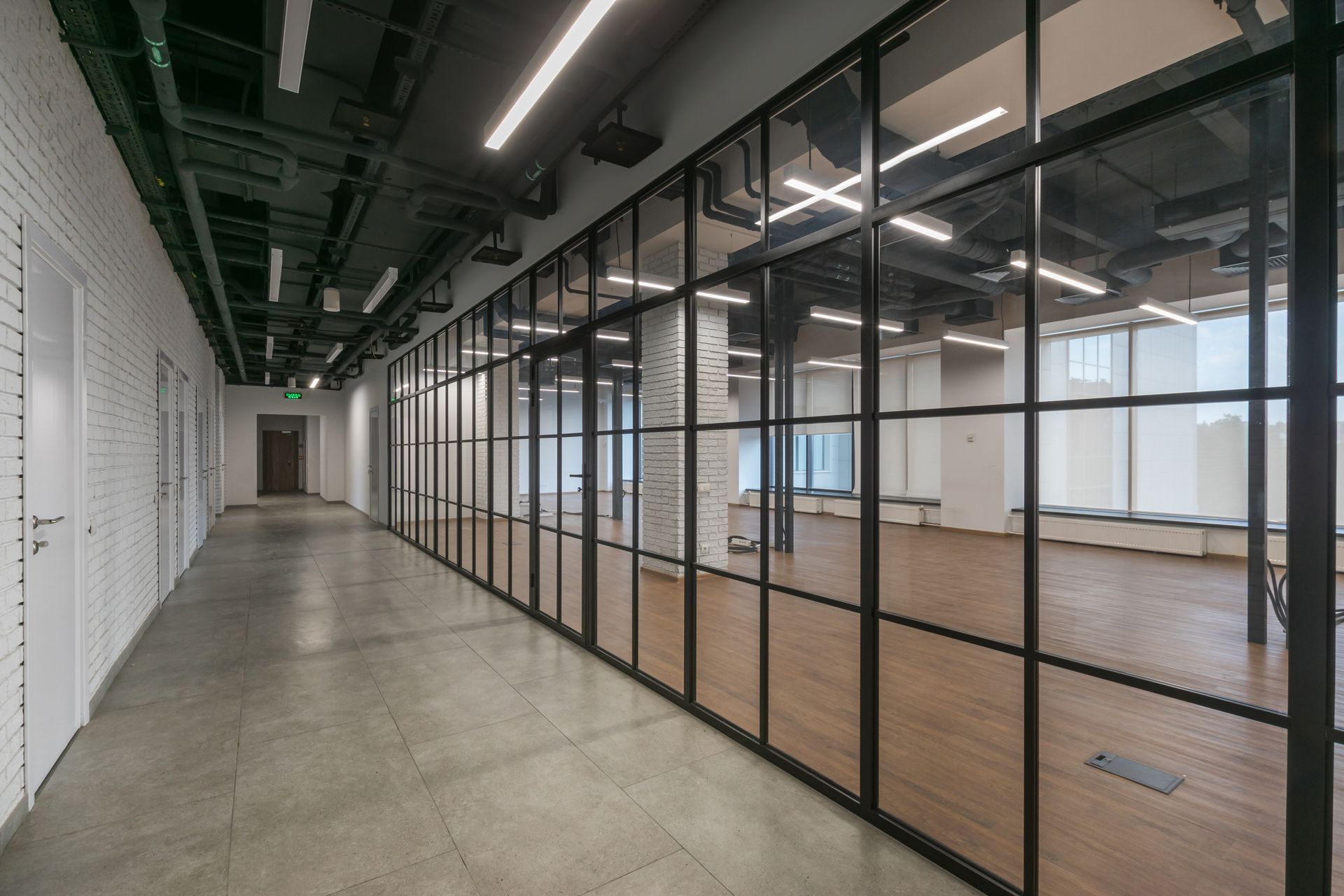 Empty office corridor beside glass-walled meeting rooms with concrete floor and overhead lights