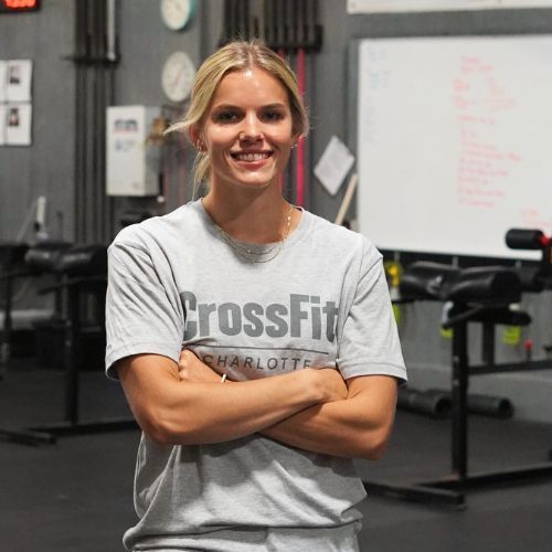 A woman wearing a grey crossfit shirt is standing in a gym with her hands on her hips.