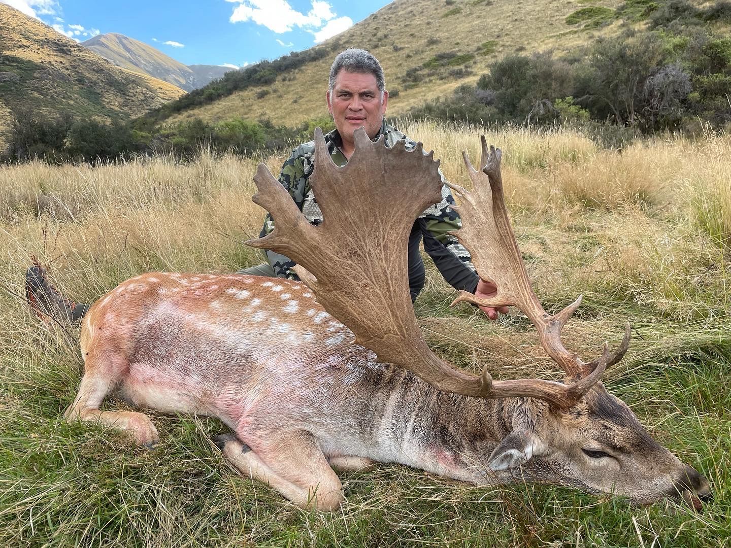 A man in camouflage poses with a large spotted fallow deer he has hunted in a grassy mountain landscape.