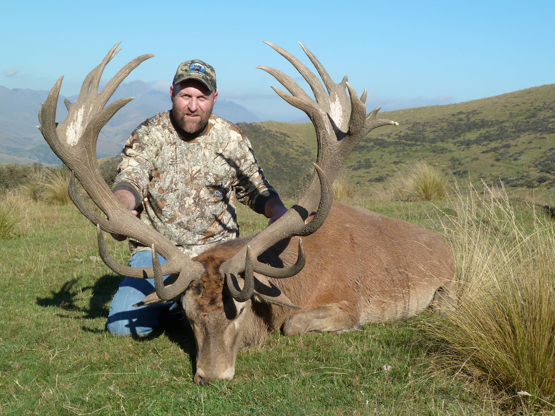 A hunter in camouflage kneels beside a large red deer with impressive antlers on a grassy hillside.