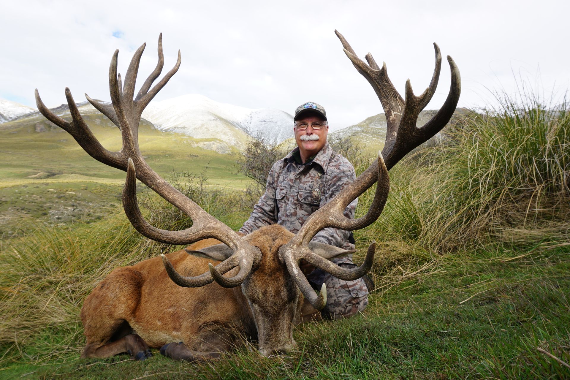 Man in camouflage kneels next to a large red deer with massive antlers in a grassy field, mountains in the background.