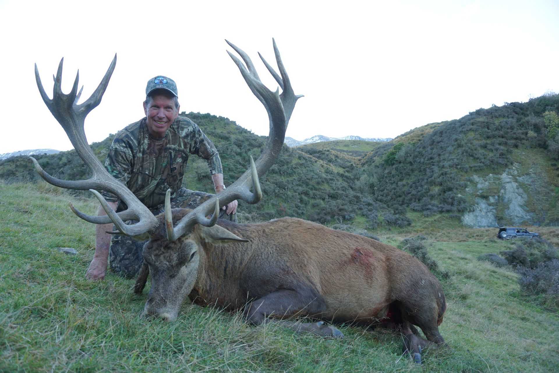 Hunter kneels next to a large red deer with impressive antlers. The hunter is smiling, in camouflage, outdoors on a grassy hill.