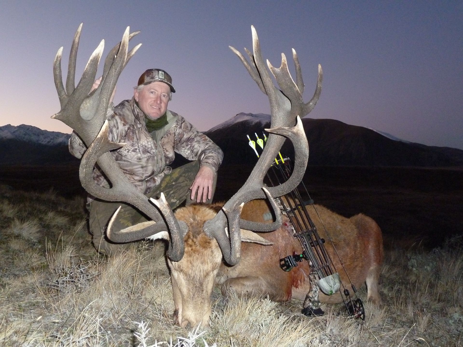 Man in camo kneeling by a large deer with impressive antlers, holding a bow, after a hunt. Mountains and dusk sky in the background.