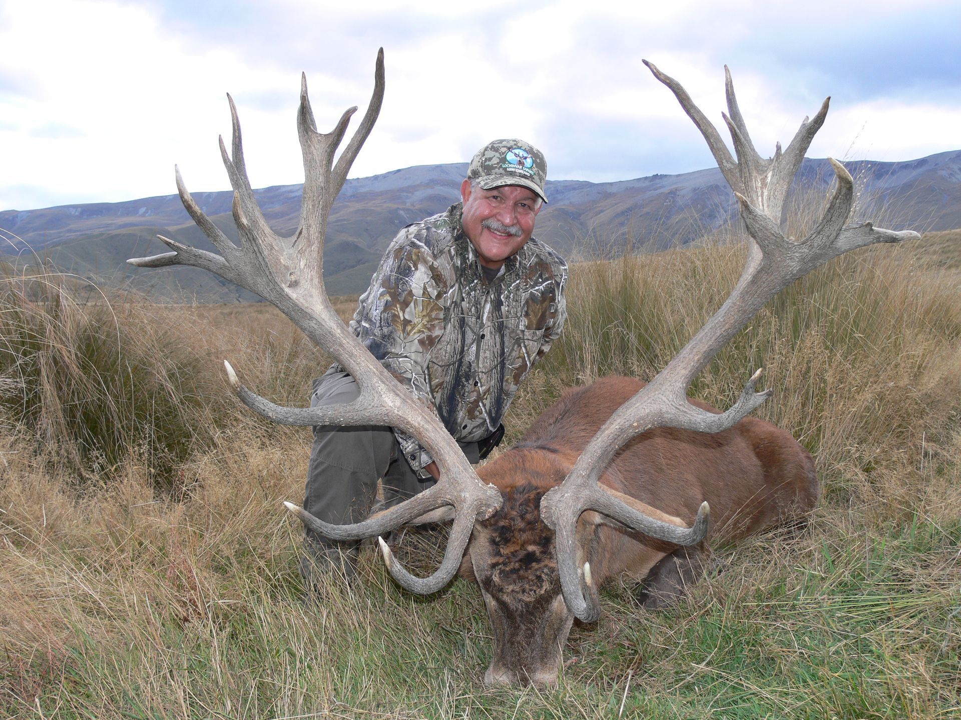 Man in camouflage kneeling next to a large, dead red deer with impressive antlers in a grassy field.