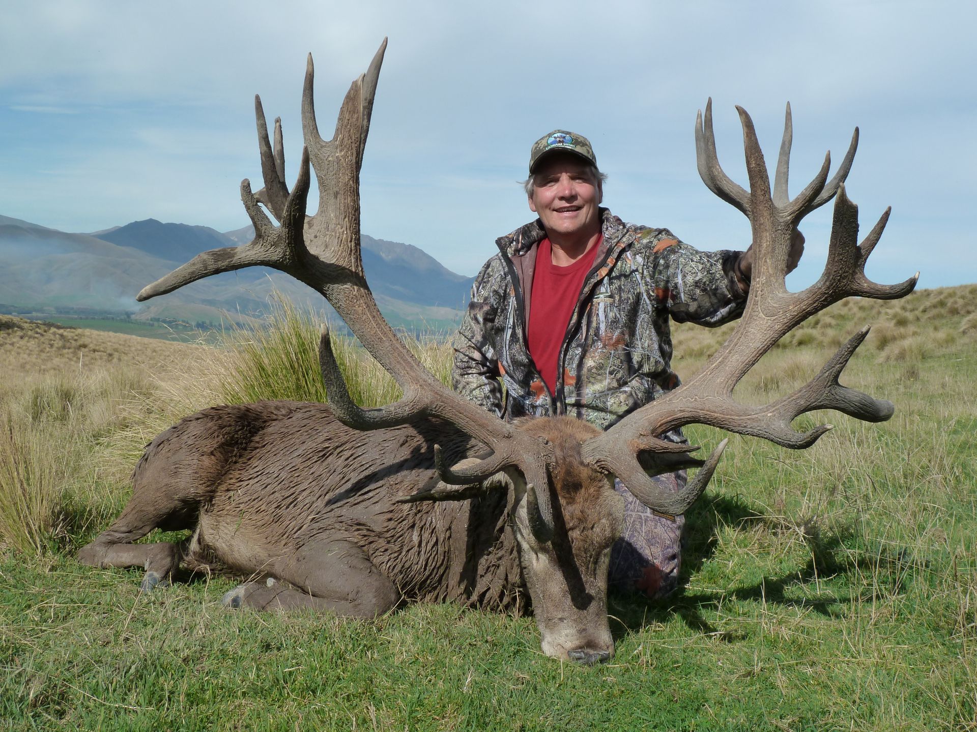 A person in camouflage kneels next to a large red deer with impressive antlers in a grassy field, mountains in the background.