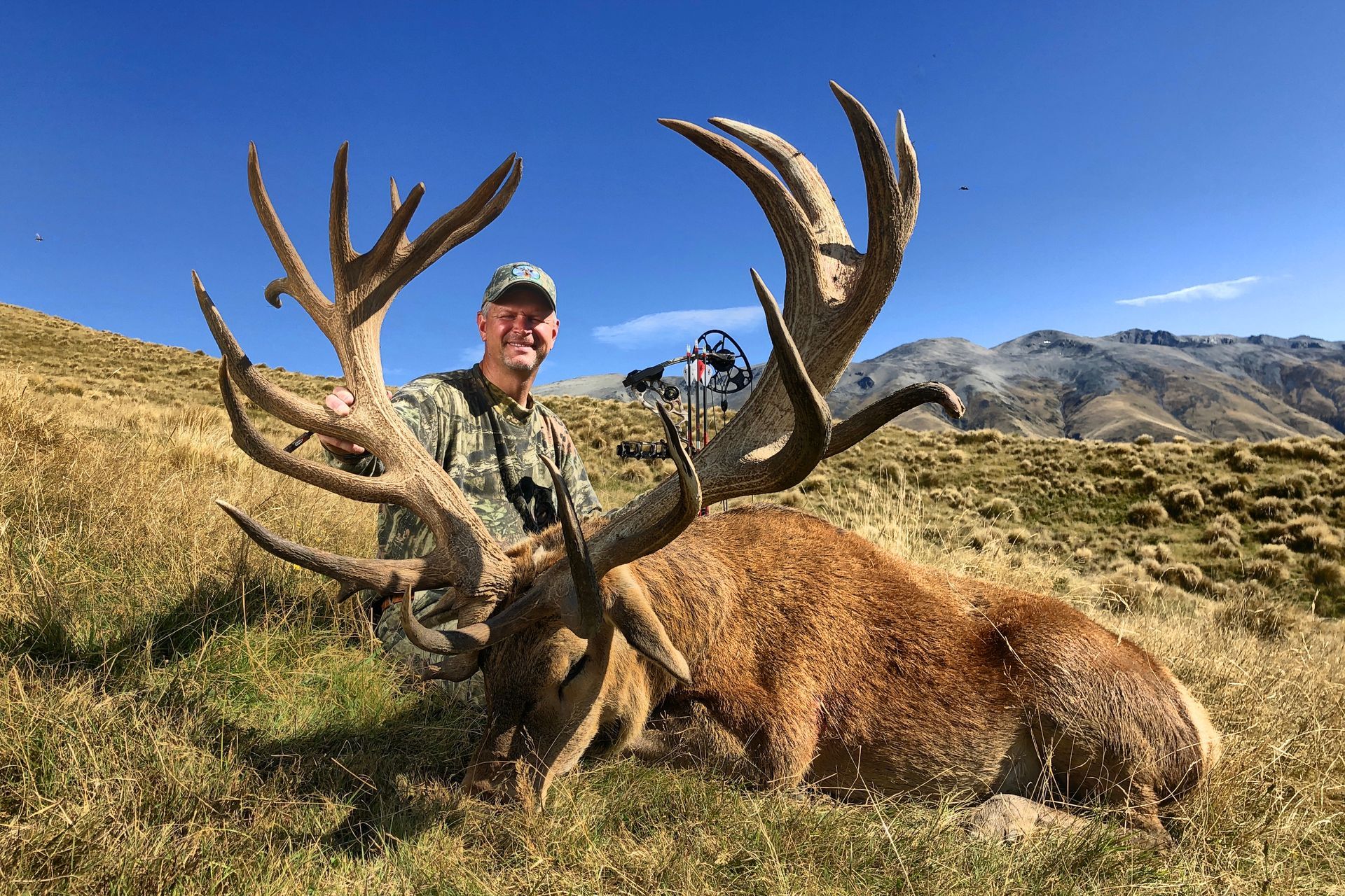 Man in camouflage smiles, posed next to a large dead elk with huge antlers, on a grassy hillside.