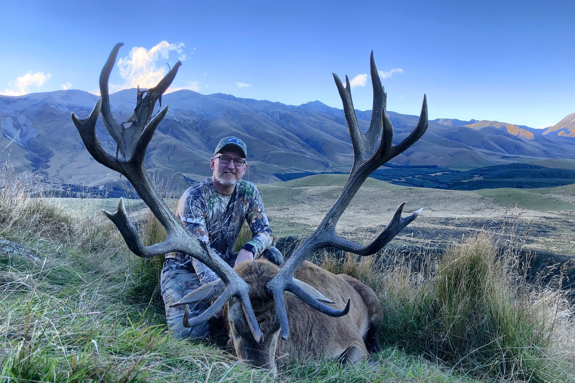 Hunter kneels beside a large red stag with impressive antlers in a mountainous, grassy landscape. The man smiles, wearing camouflage.