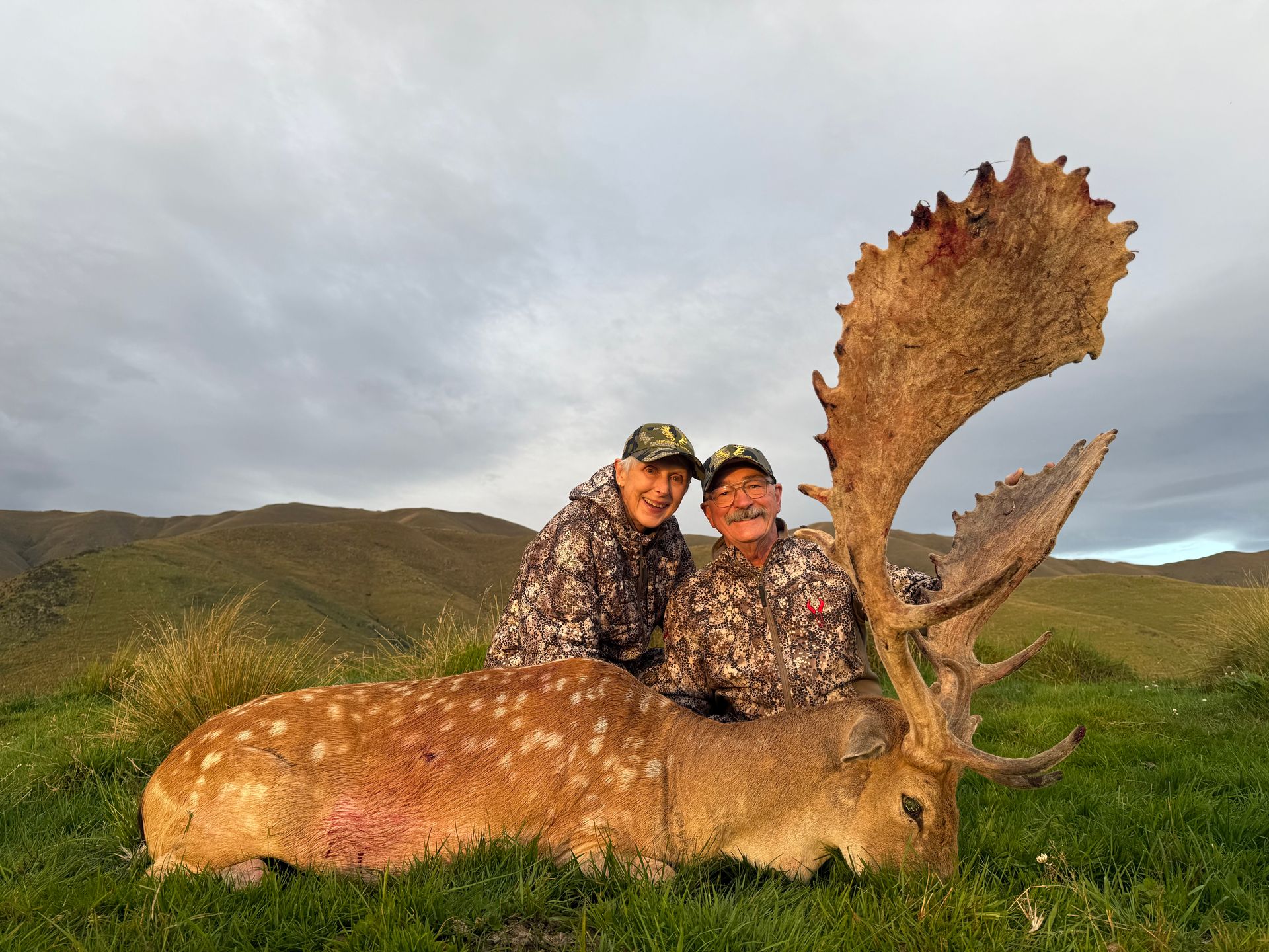 Two people in camouflage smile next to a harvested fallow deer with large antlers on a grassy hillside.