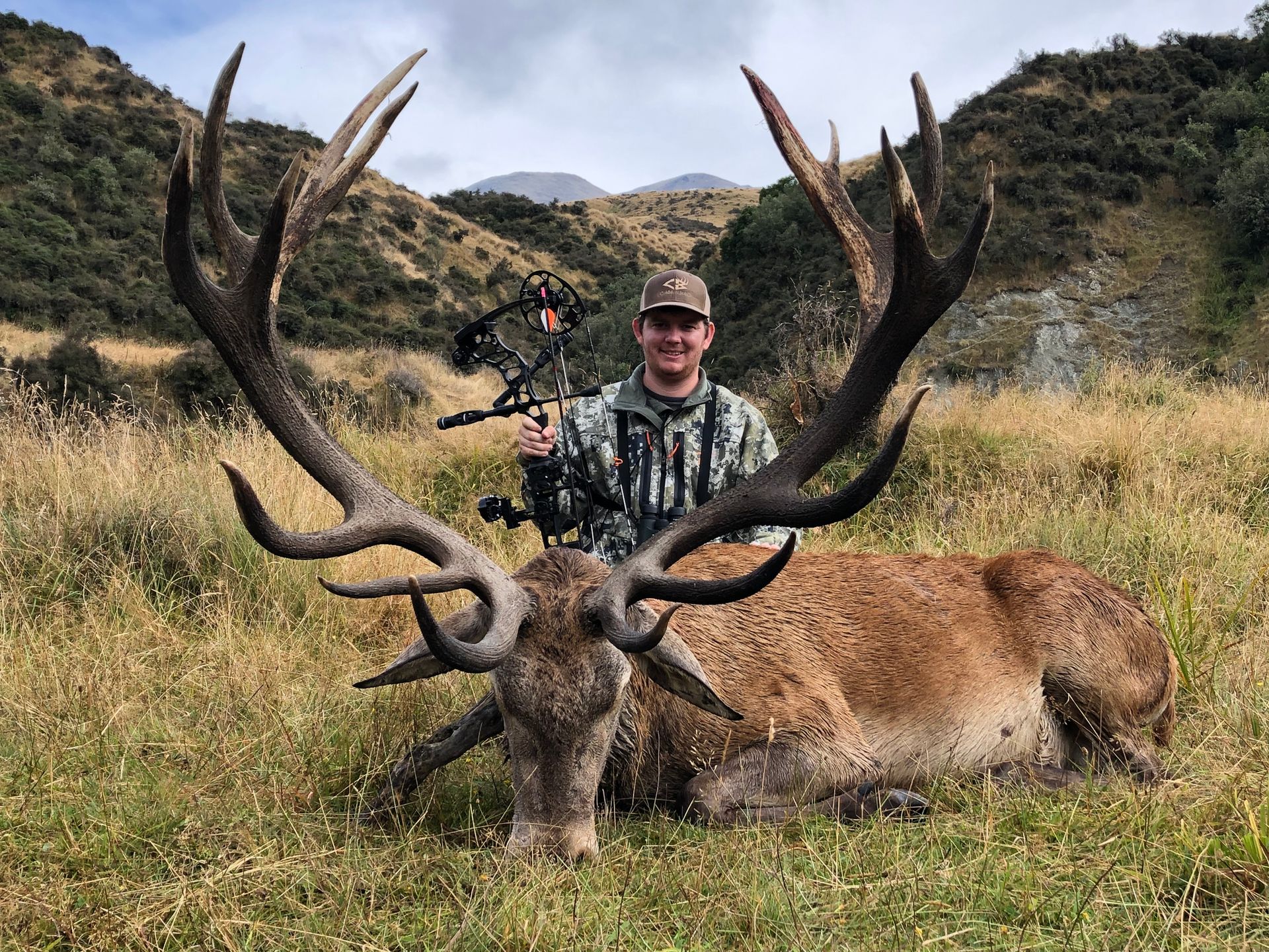 Man in camouflage poses with a large dead red deer in a grassy field, holding a bow and arrow. Mountains are in the background.