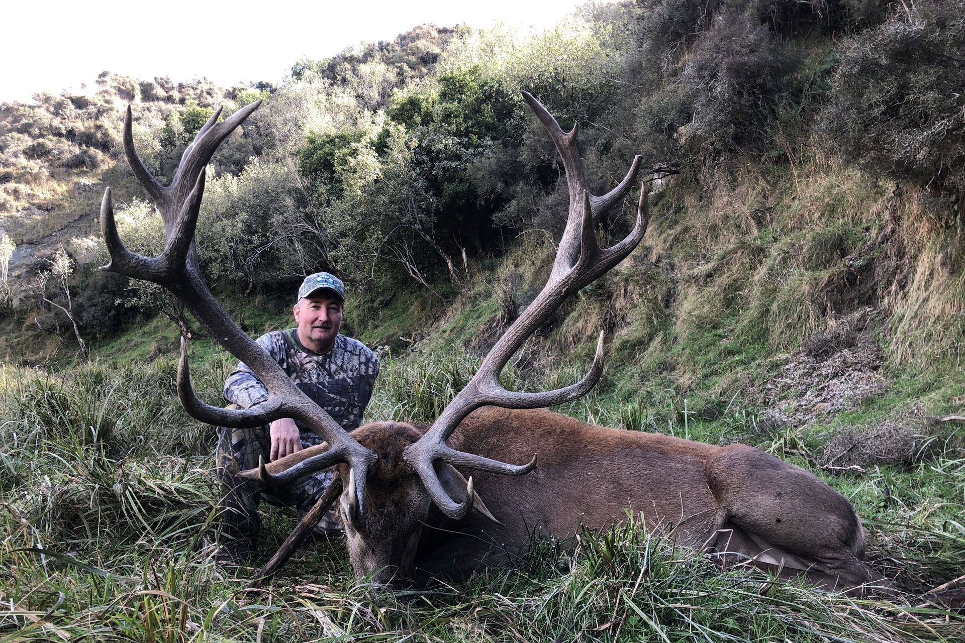 Man kneeling beside a large red stag with massive antlers, in a grassy, natural setting.