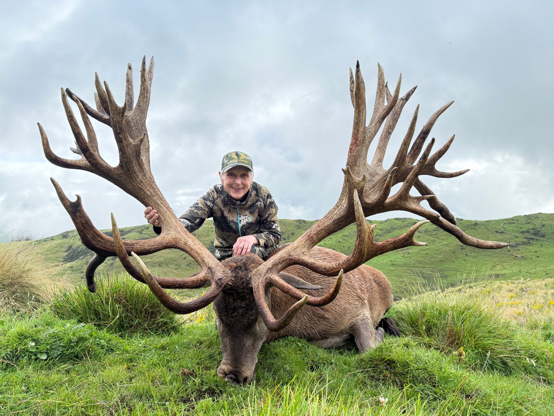 Person in camouflage next to a large dead stag with enormous antlers, outdoors in a grassy field, smiling.