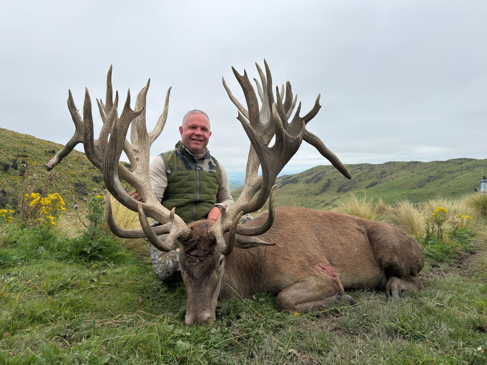Man smiles, sitting beside a large red deer with impressive antlers, in a grassy, outdoor setting.