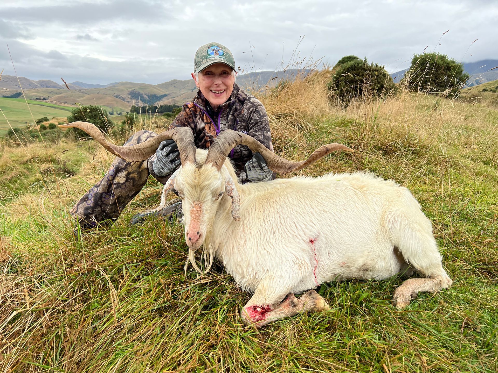 A person in camouflage gear poses with a white goat with large horns in a grassy, hilly landscape. The goat has a bloodied wound.