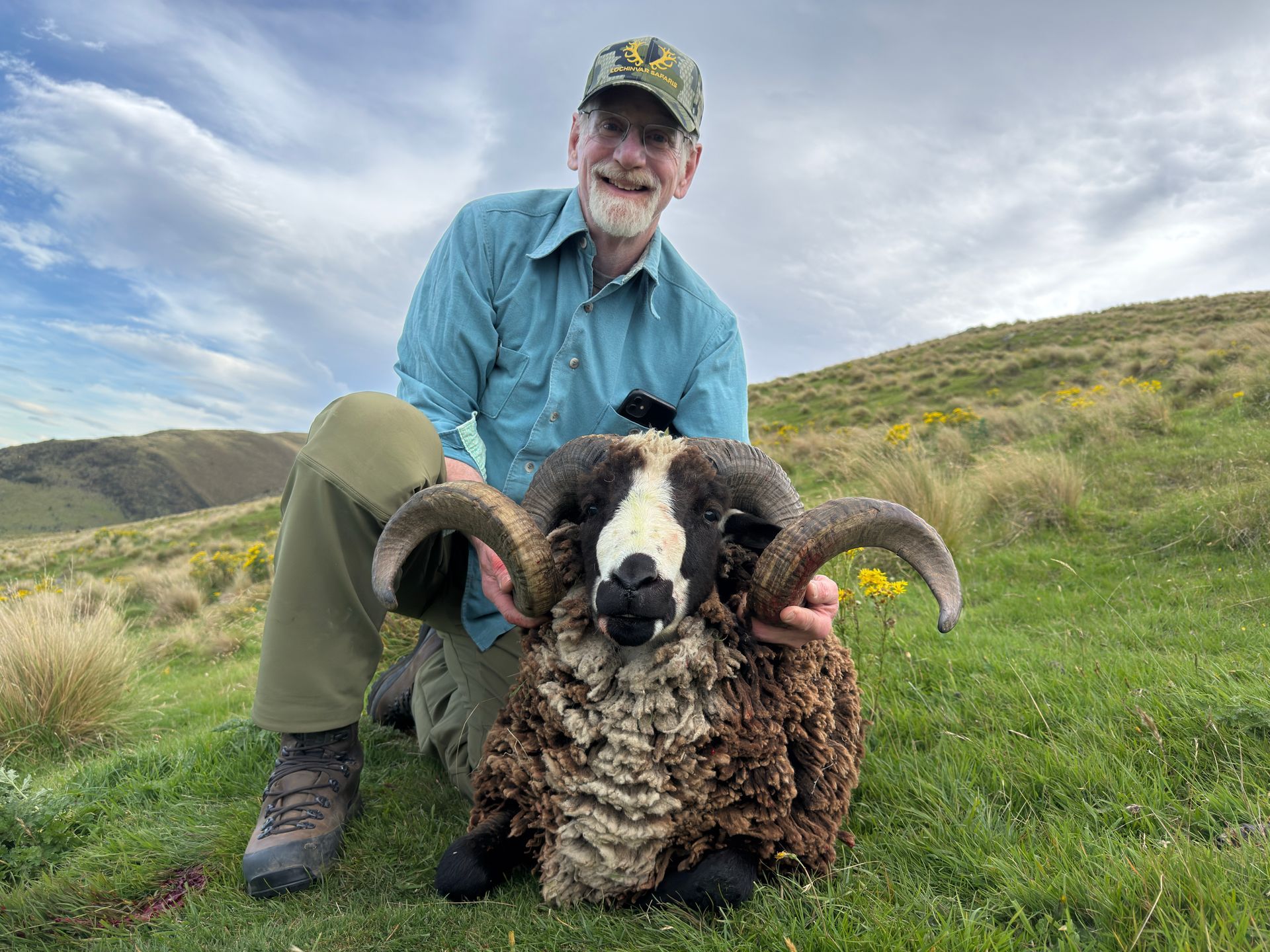 Man kneels beside a large ram with curled horns, smiling for the camera in a grassy field.