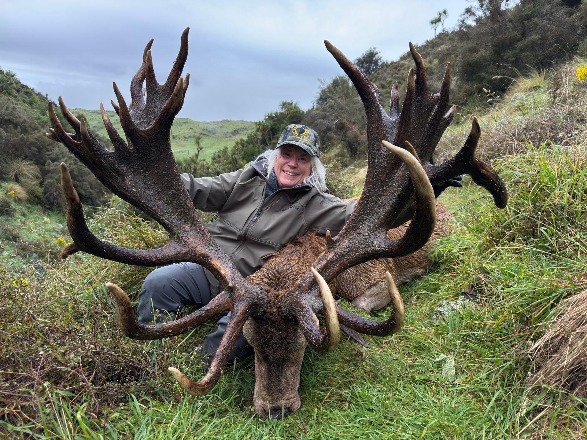 Man in camouflage hat kneels beside a large stag with huge antlers. He is smiling, and they are in a grassy field.