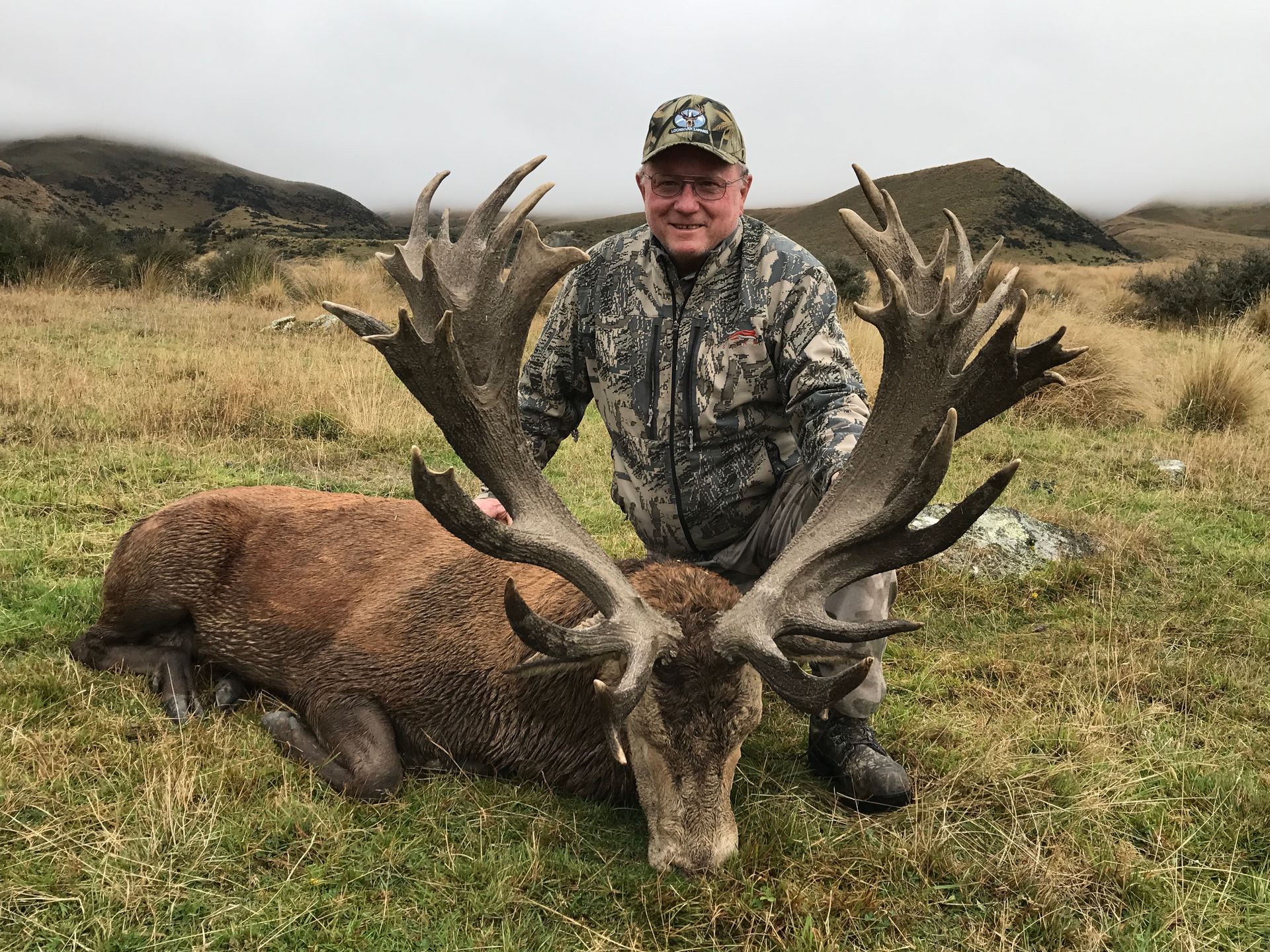 A man in camouflage stands next to a large, dead deer with large antlers in a grassy field, mountainous background.