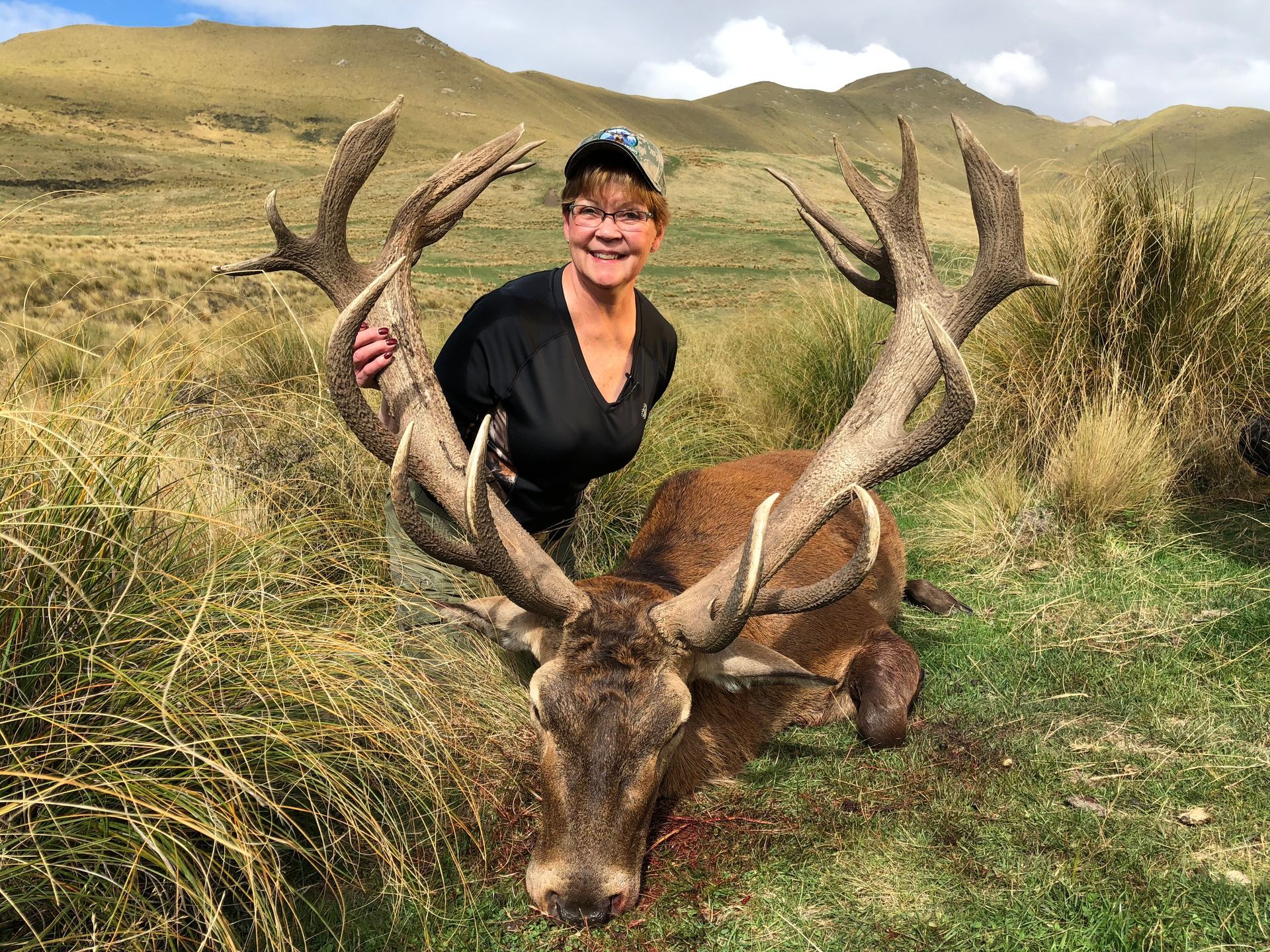 Woman smiling, poses with a large dead deer with impressive antlers in a grassy field.
