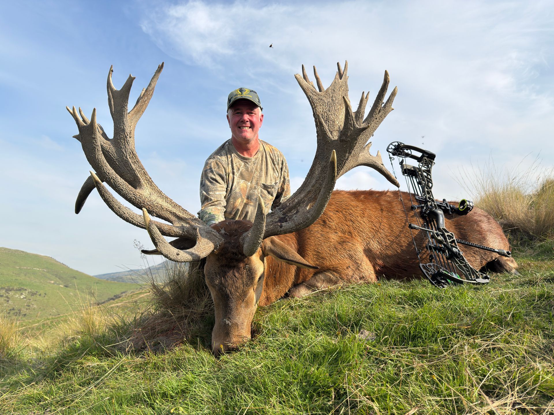 Man in camouflage smiling, posing with a large, dead red stag, next to a bow, in a grassy field with hills in the background.