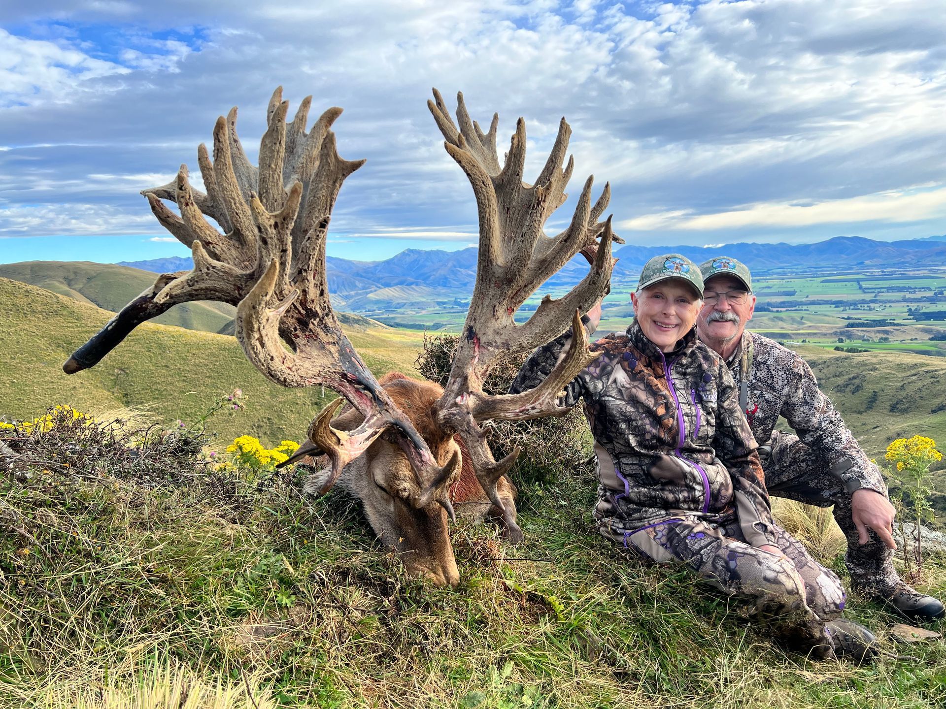 Two people in camouflage pose beside a large deer with impressive antlers on a hillside with a scenic vista.