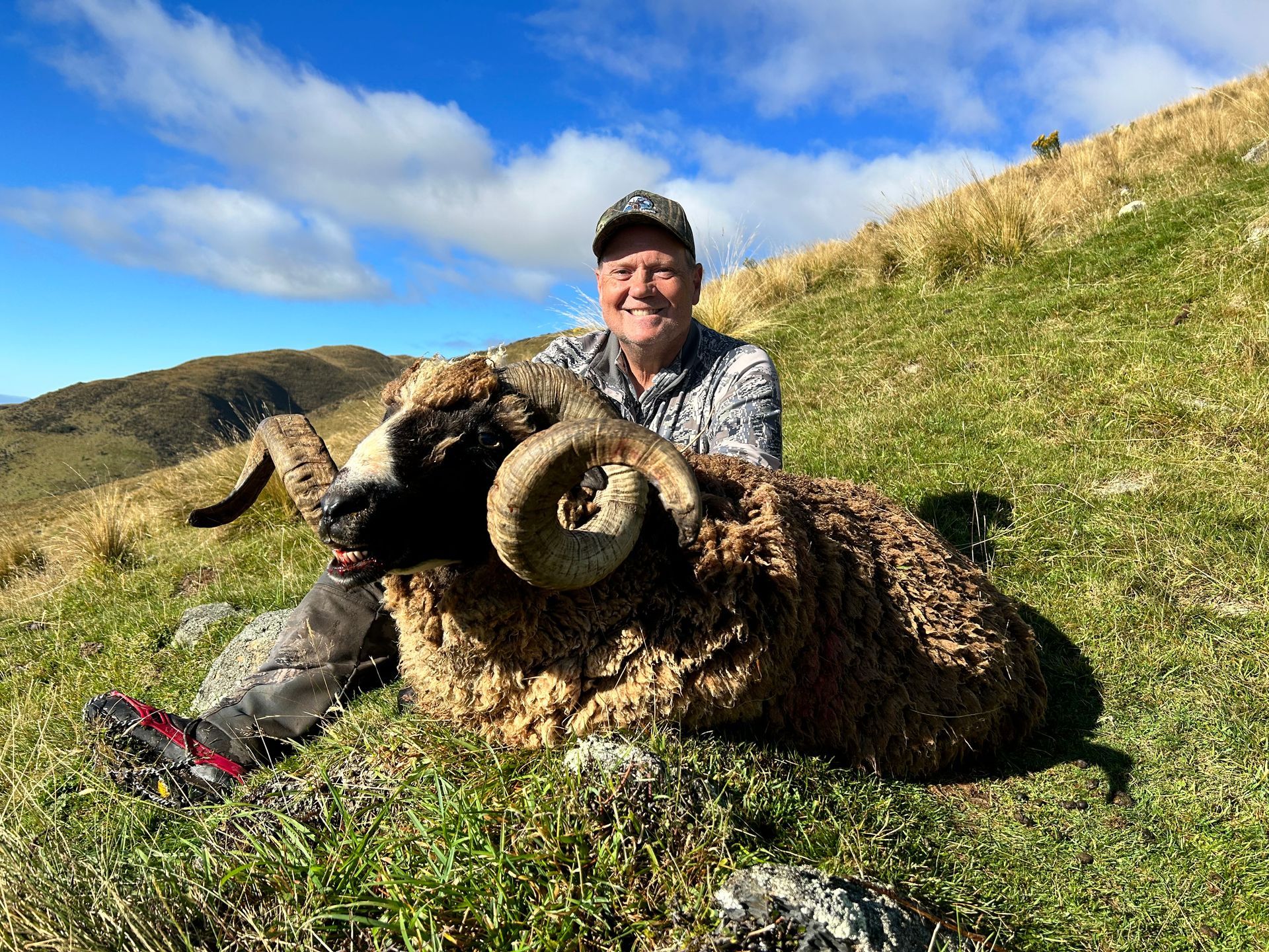 A smiling man in camouflage gear sits with a large, curled-horn ram on a grassy hillside.