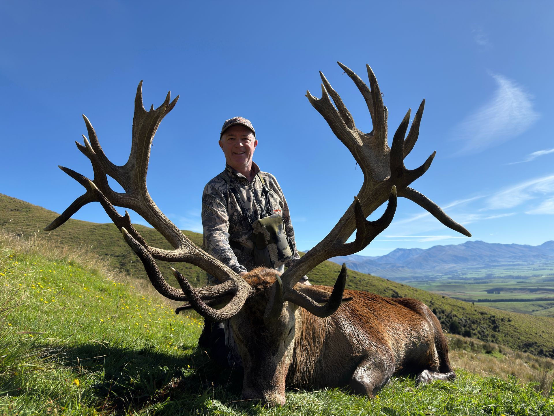 Hunter stands with a large red deer stag he has killed, with massive antlers, in a grassy field under a blue sky.