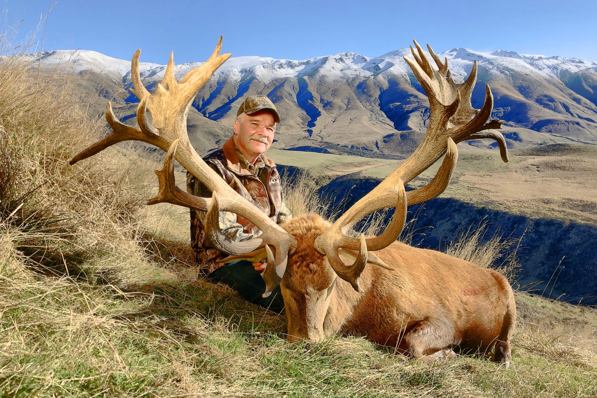 Man in camouflage kneels beside a large red deer with impressive antlers in a mountainous, sunny setting.