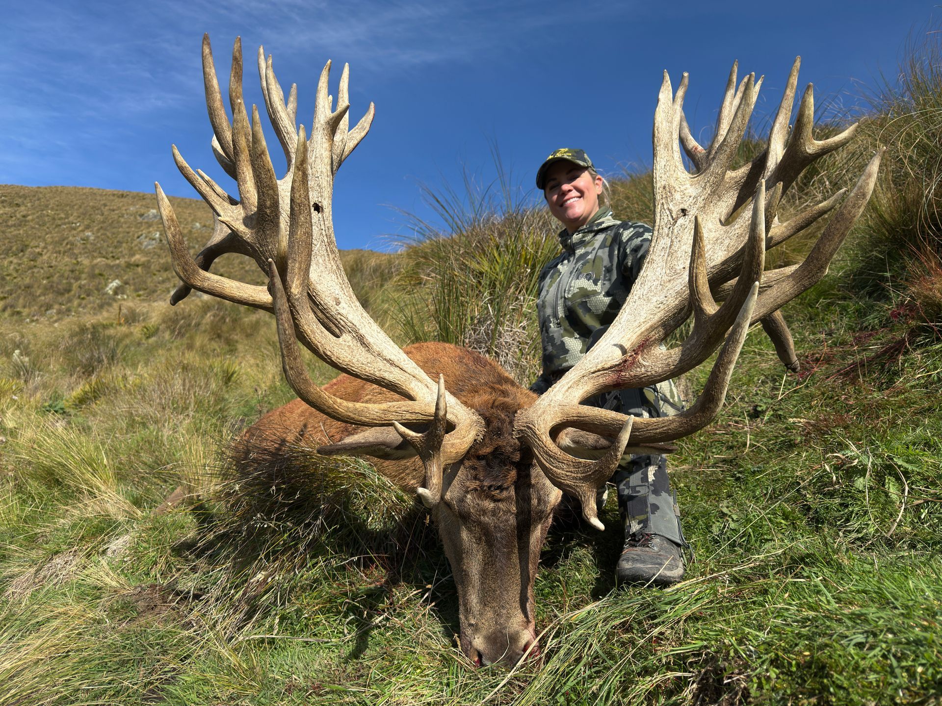 Hunter smiles, posing with a large-antlered red deer it has killed on a grassy hillside under a blue sky.