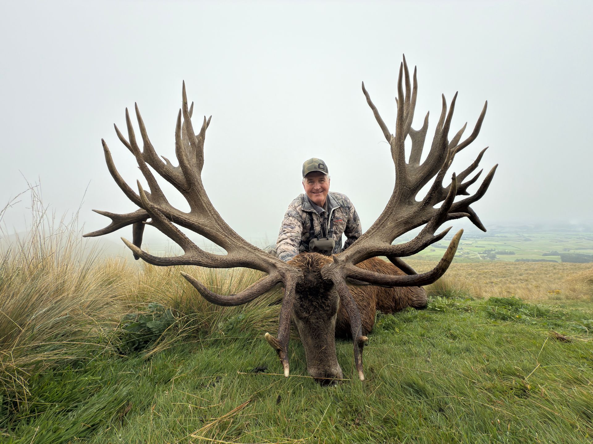 Hunter poses with a large red deer stag with massive antlers, on a grassy hillside under an overcast sky.