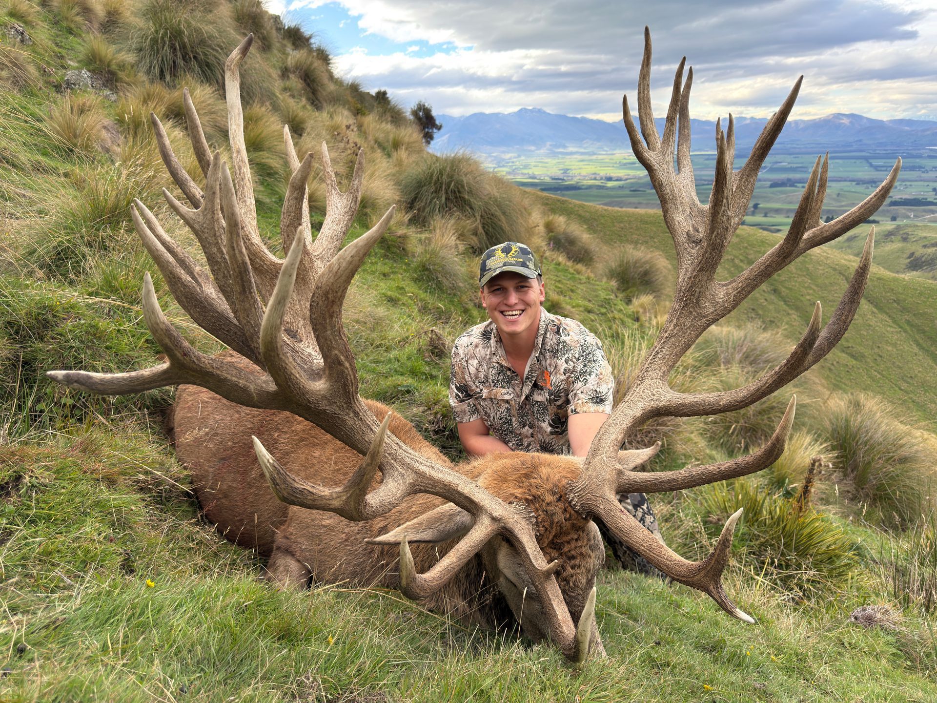 A smiling man in camouflage sits next to a large-antlered red deer on a grassy hillside. Mountains are in the background.