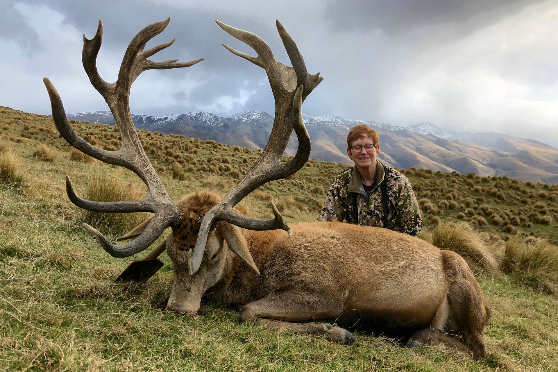 A hunter poses with a large dead stag in a grassy field with mountains in the background. The stag has massive antlers.