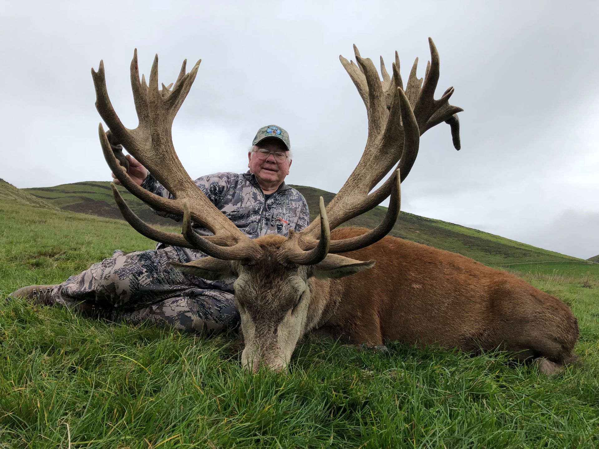 Man in camouflage sitting next to a large dead red deer on a grassy hillside. Deer has large antlers.