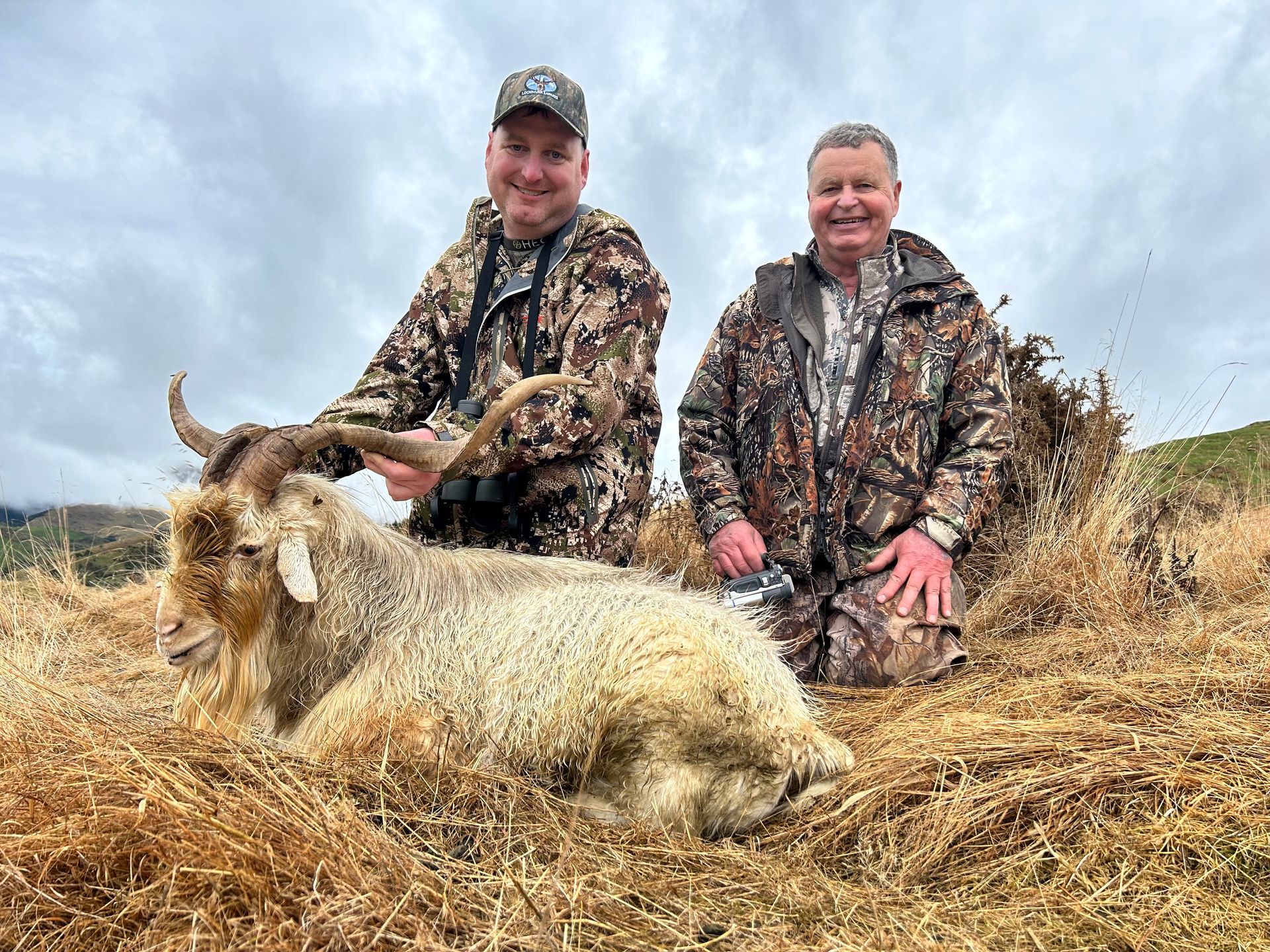 Two men in camouflage pose with a large, shaggy goat they appear to have hunted, outdoors on a cloudy day.