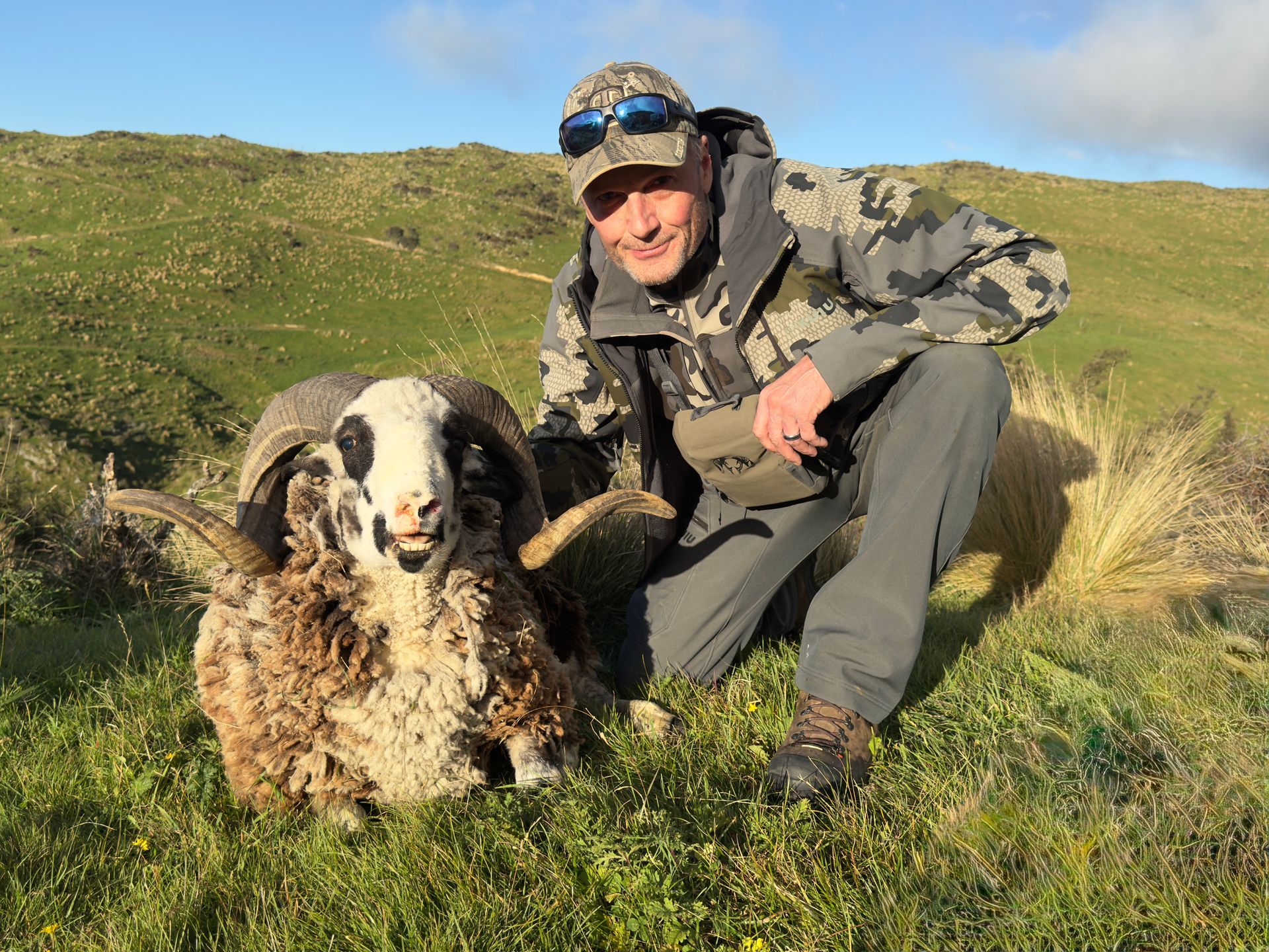 Man kneels beside a large ram with curly horns, in grassy hills. The man wears camouflage and smiles.