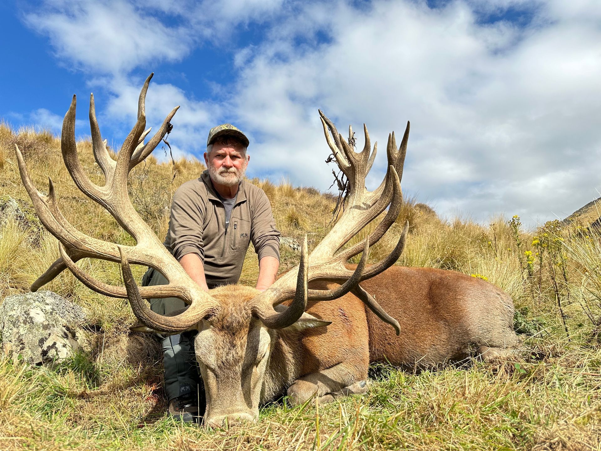 Man poses with a large dead red deer, its antlers sprawling. Grassy hillside setting under a partly cloudy sky.