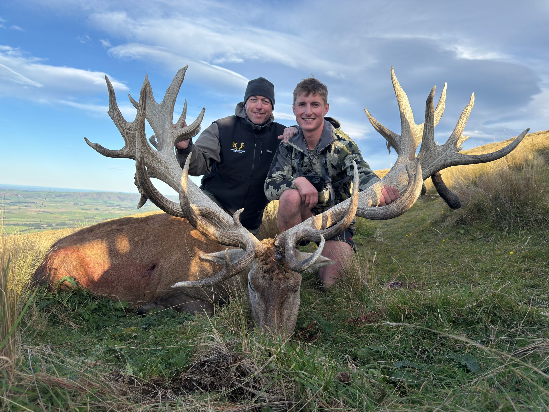 Two men posing with a large red deer stag with large antlers on a grassy hillside.