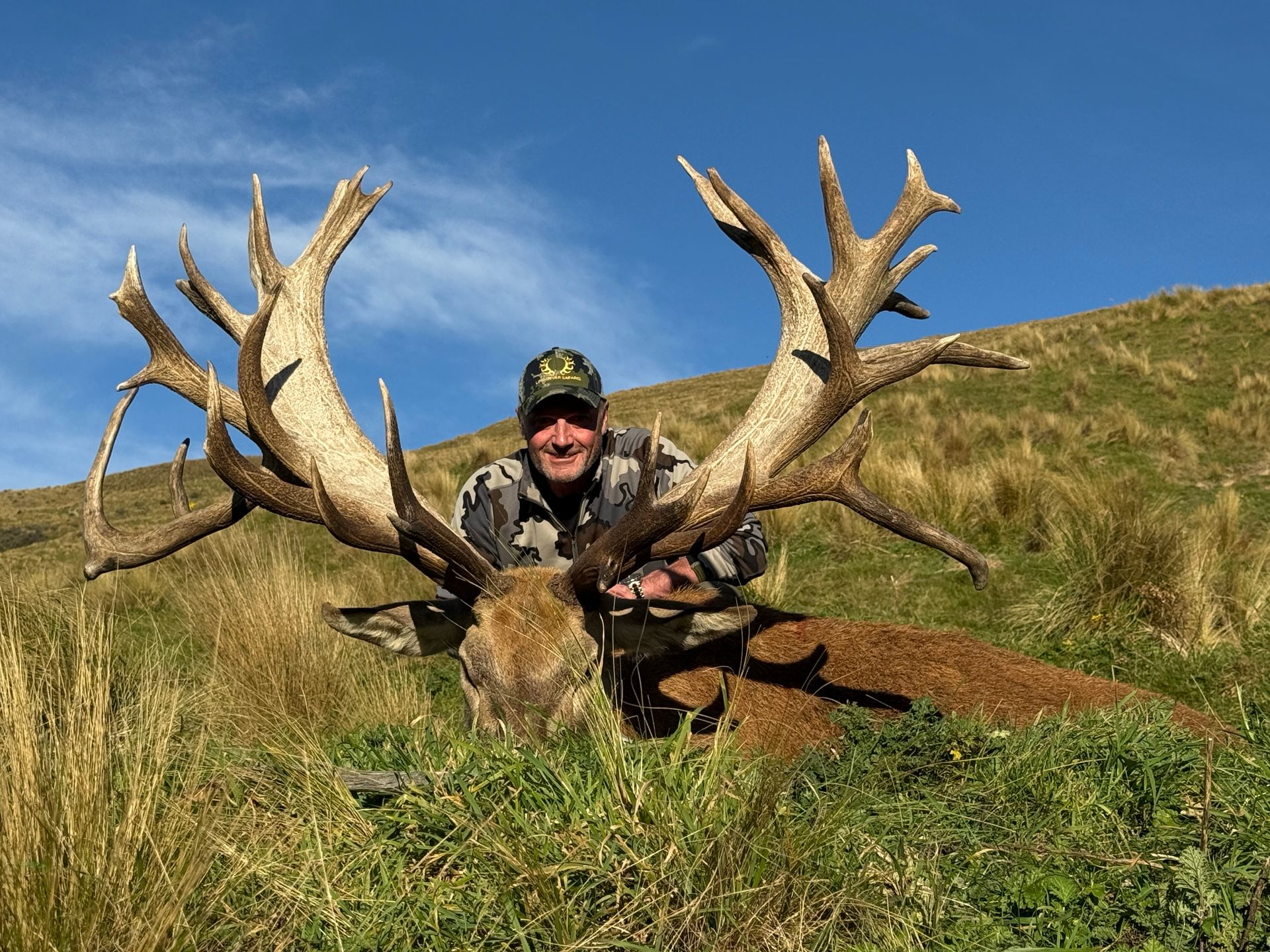 Man in camouflage with large deer, posing on grassy hillside under blue sky. Deer has large antlers.