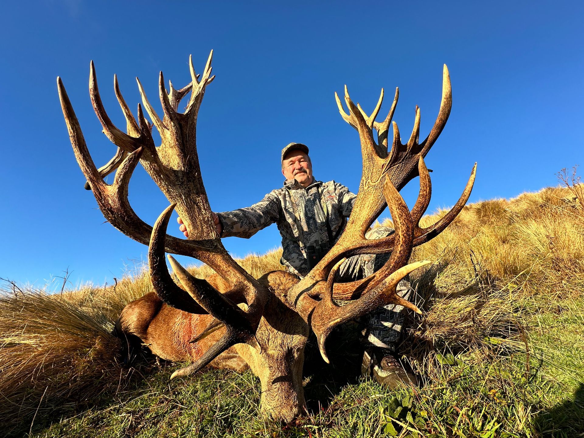 Hunter smiling, posing behind large red deer stag with massive antlers in a grassy field against a blue sky.