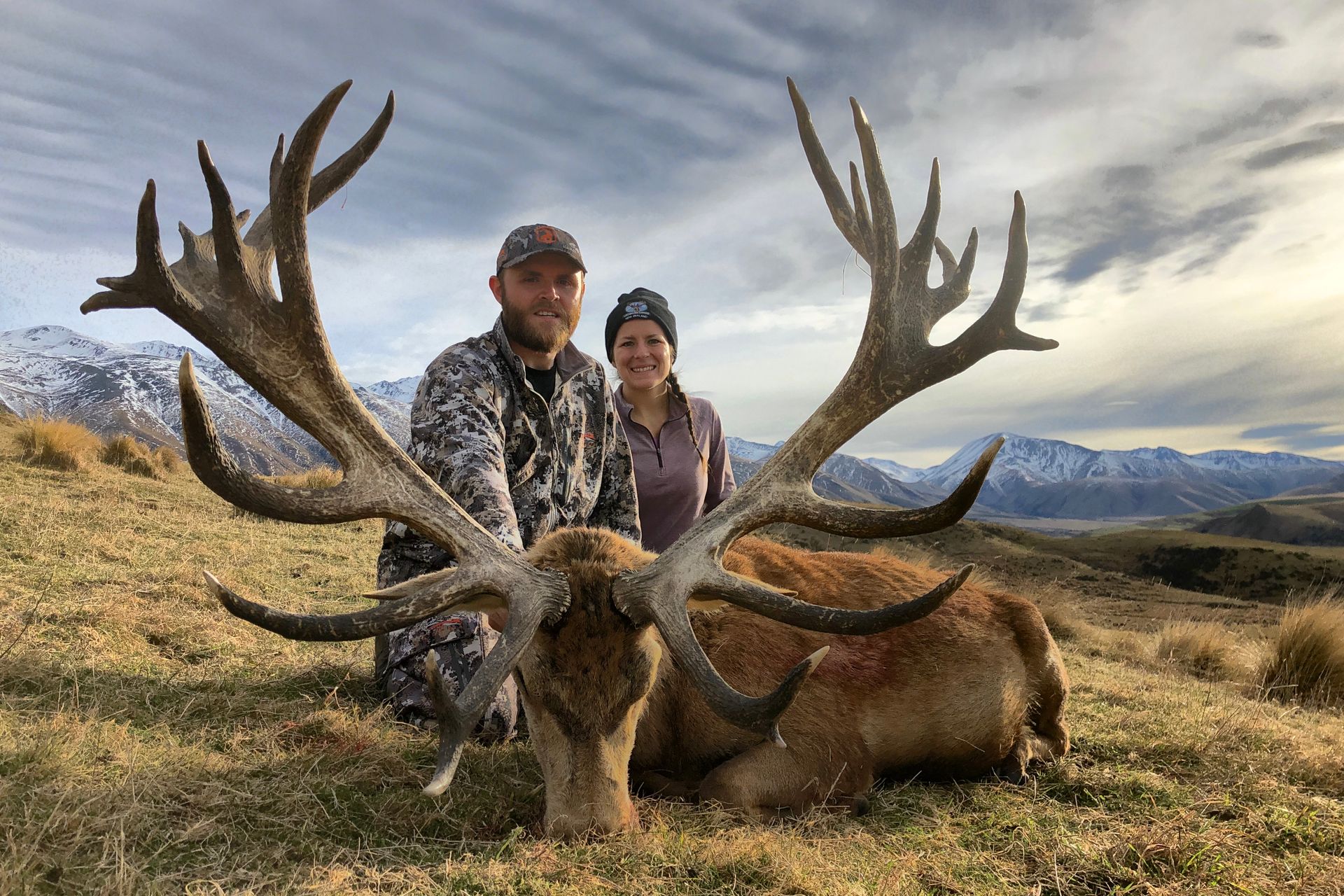 A man and woman pose with a large, harvested deer with impressive antlers in a grassy mountain landscape.