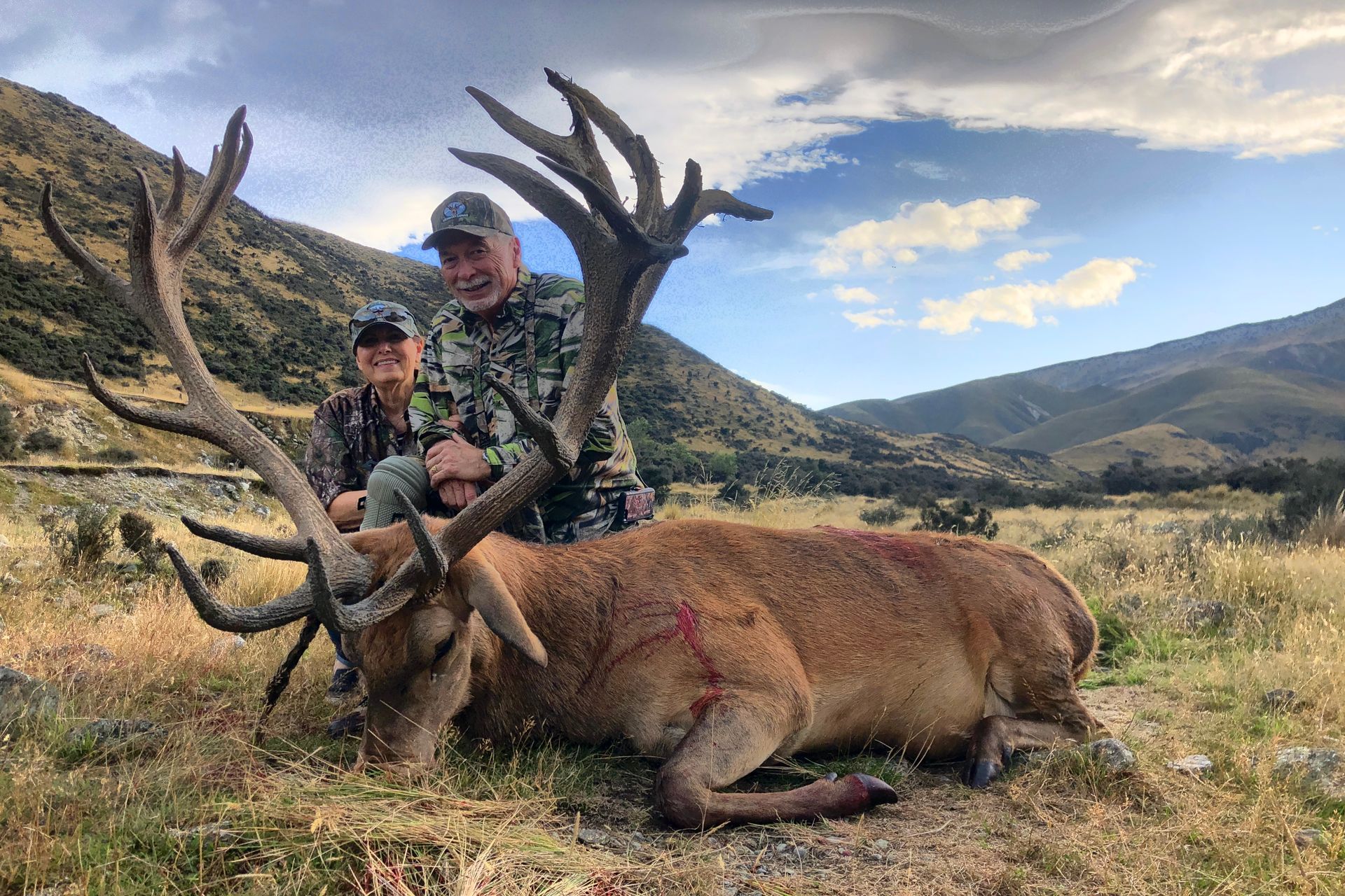 Two hunters pose with a large red deer stag in a grassy mountainous setting. The deer has large antlers and appears deceased.