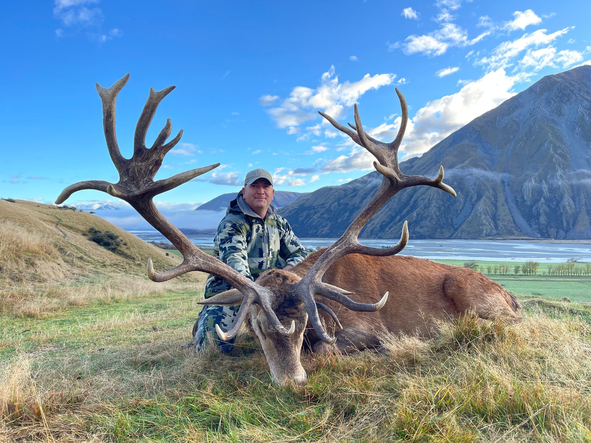 Hunter kneeling beside a large red deer with impressive antlers, posing on a grassy hillside with mountains and a river in the background.