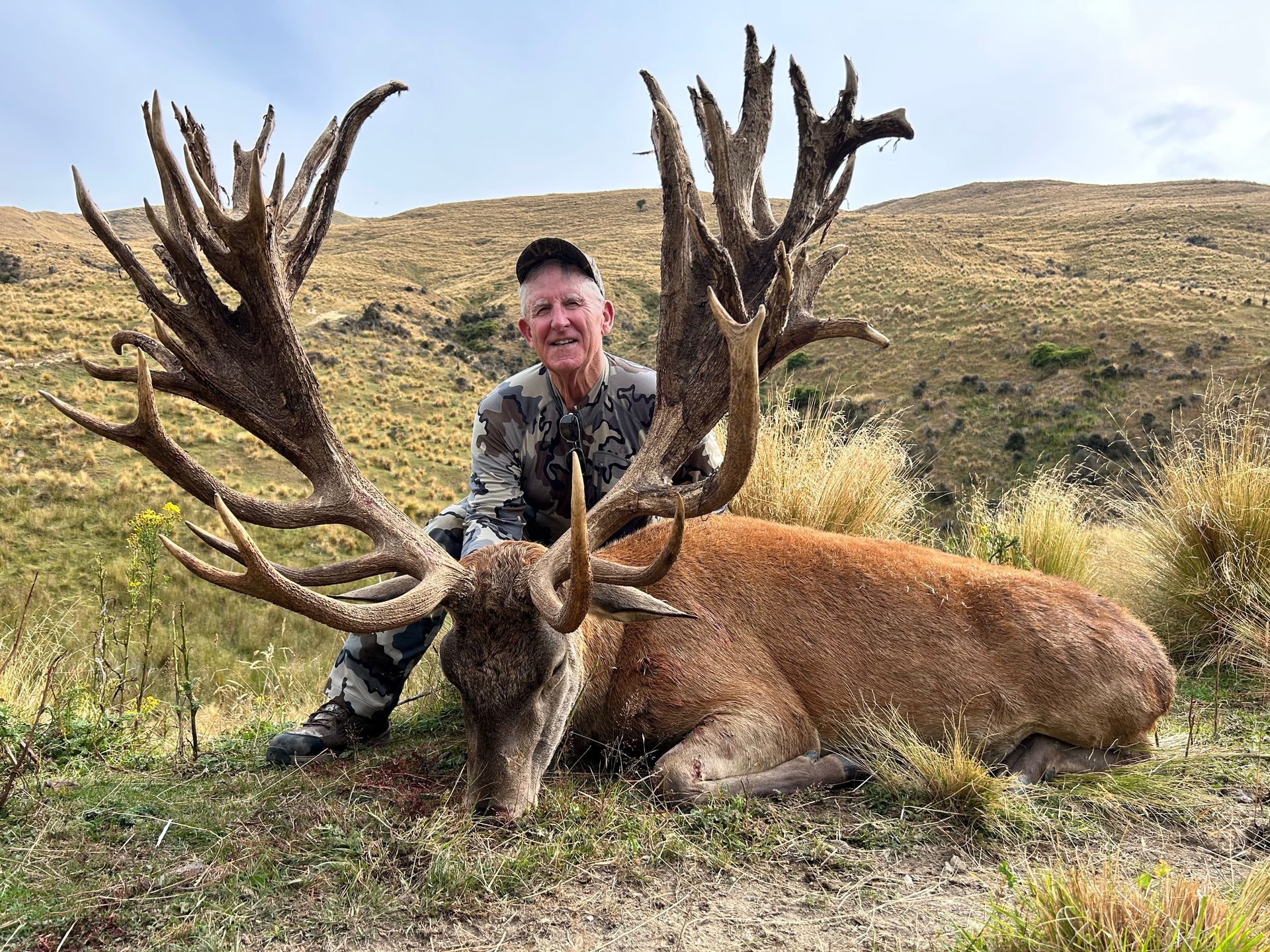 Man in camouflage kneels next to a large red deer with massive antlers, posing for a photo in a grassy, hilly setting.