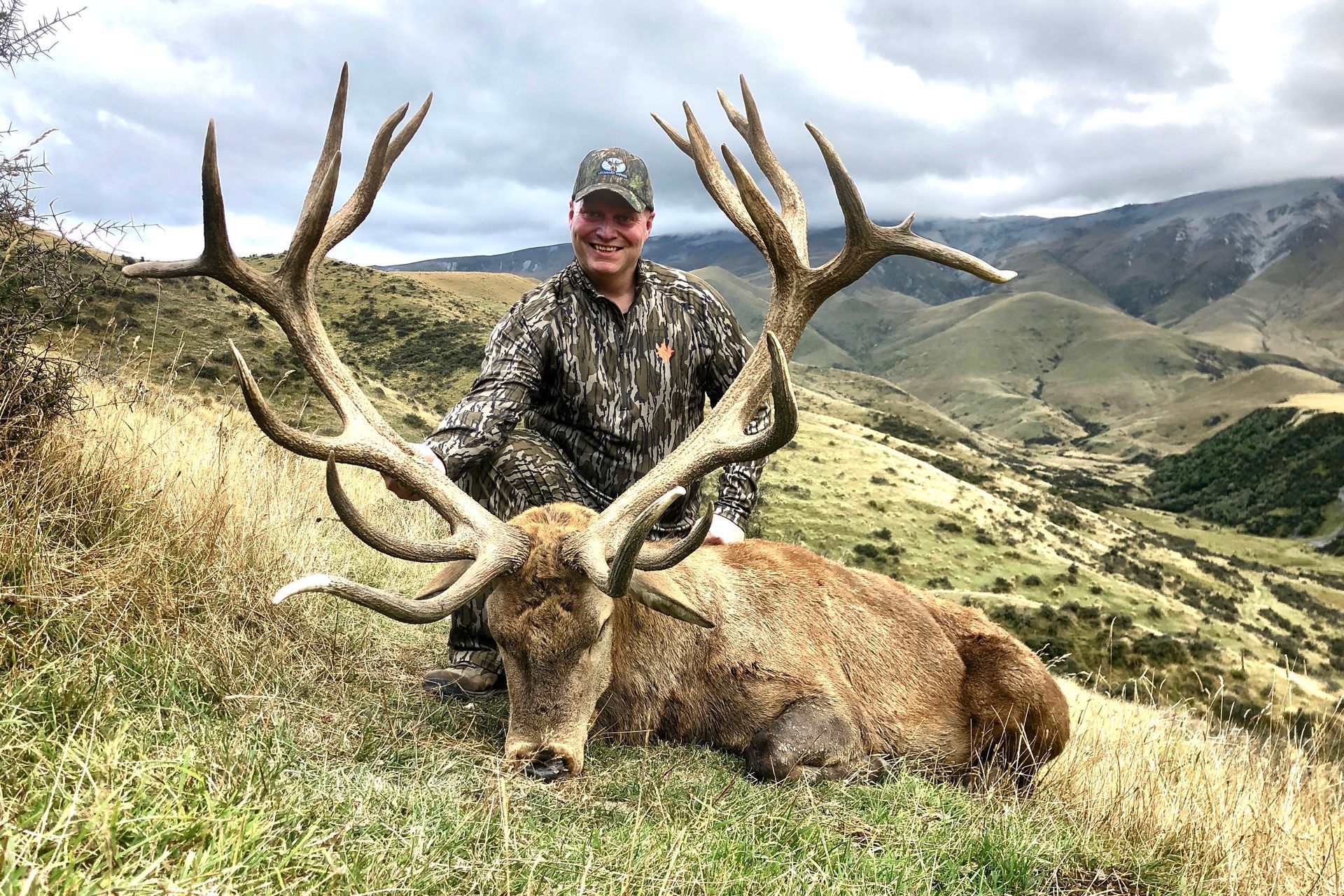 Man in camouflage kneels next to a large red deer with impressive antlers in a grassy, mountainous landscape. The man smiles.